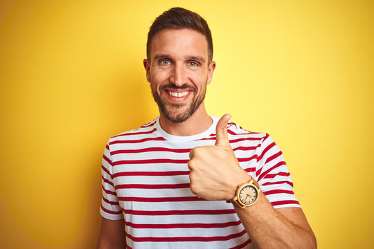 Young handsome man wearing casual red striped t-shirt over yellow isolated background doing happy thumbs up gesture with hand. Approving expression looking at the camera showing success.