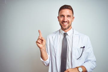 Young handsome doctor man wearing white profressional coat over isolated background with a big smile on face, pointing with hand and finger to the side looking at the camera.