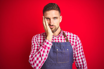 Young handsome man wearing shop owner apron uniform over red isolated background thinking looking tired and bored with depression problems with crossed arms.