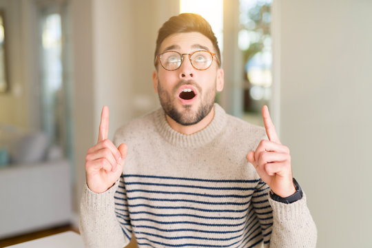 Young handsome man wearing glasses at home amazed and surprised looking up and pointing with fingers and raised arms.