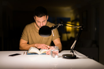 Young handsome man studying at home, reading a book at night
