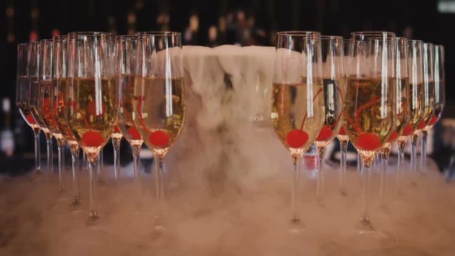 Glasses with alcohol, champagne, wine and cherries below stand in a row in a nightclub bar. White cold smoke spreads out from a bucket of dry ice. Cooking chilled alcoholic cocktails.