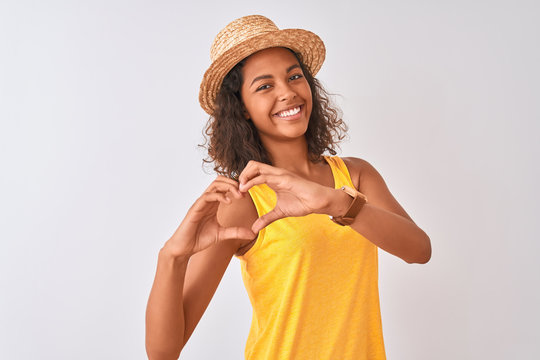 Young brazilian woman wearing yellow t-shirt and summer hat over isolated white background smiling in love showing heart symbol and shape with hands. Romantic concept.
