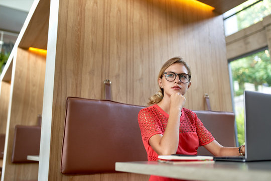 Closeup lifestyle portrait of young pensive blonde caucasian millennial blogger wearing glasses taking notes on a laptop in a bright trendy restaurant