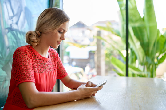 Lifestyle shot of young blonde caucasian natural millennial woman working on cellphone at a modern tropical and bright office with plants