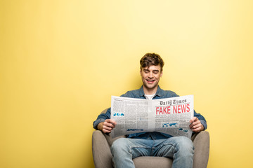 smiling young man sitting in armchair and reading newspaper with fake news on yellow background