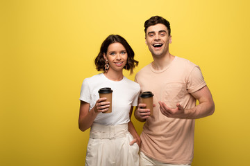 cheerful man and woman holding coffee to go while smiling a camera on yellow background