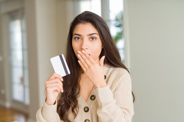 Young woman holding credit card as payment cover mouth with hand shocked with shame for mistake, expression of fear, scared in silence, secret concept