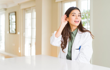Young woman wearing medical coat at the clinic as therapist or doctor smiling with hand over ear...