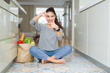 Young woman sitting on the kitchen floor with a paper bag full of fresh groceries smiling making frame with hands and fingers with happy face. Creativity and photography concept.