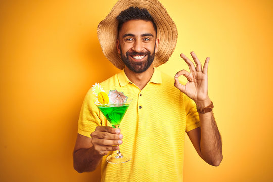Indian man on vacation drinking cocktail wearing summer hat over isolated yellow background doing ok sign with fingers, excellent symbol