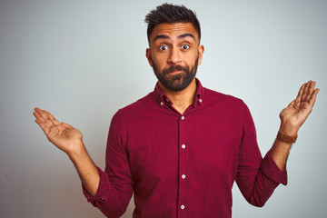 Young indian man wearing red elegant shirt standing over isolated grey background clueless and...