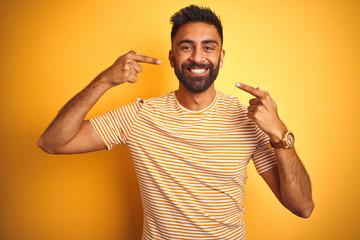Young indian man wearing t-shirt standing over isolated yellow background smiling cheerful showing and pointing with fingers teeth and mouth. Dental health concept.