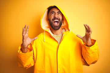 Young indian man wearing raincoat with hood standing over isolated yellow background crazy and mad...