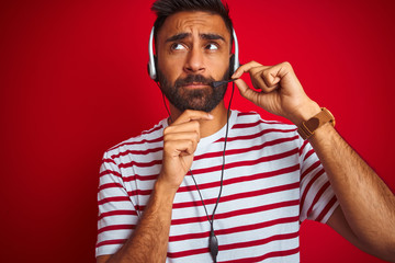 Young indian call center agent man using headset over isolated red background serious face thinking...