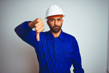 Handsome indian worker man wearing uniform and helmet over isolated white background looking unhappy and angry showing rejection and negative with thumbs down gesture. Bad expression.