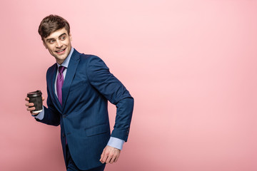 cheerful businessman looking back while holding paper cup on pink background