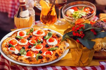 Italian pizza with tomatoes, mozzarella cheese, basil, black olives. In background, wine and highlight of light at sunset