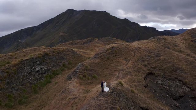 Cinematic Slow Motion of Married Couple in Wedding Suits Kissing on Top of Hill on Strong Wind. Helicopter Aerial