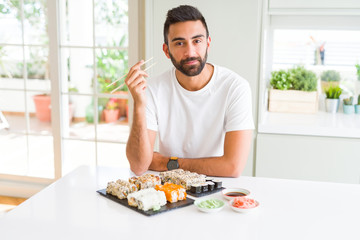 Handsome hispanic man eating asian sushi using chopsticks with a confident expression on smart face thinking serious