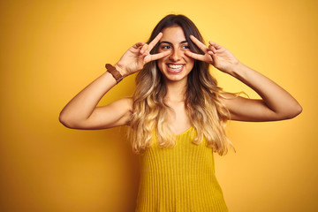 Young beautiful woman wearing t-shirt over yellow isolated background Doing peace symbol with fingers over face, smiling cheerful showing victory