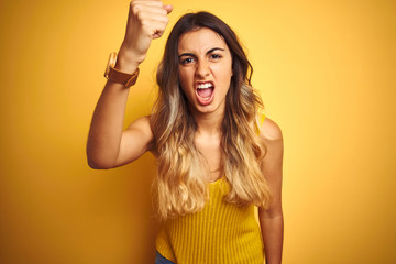 Obraz na płótnie Canvas Young beautiful woman wearing t-shirt over yellow isolated background angry and mad raising fist frustrated and furious while shouting with anger. Rage and aggressive concept.