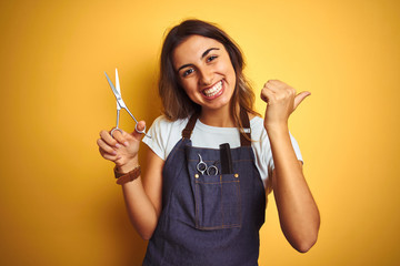 Young beautiful hairdresser woman holding scissors over yellow isolated background pointing and showing with thumb up to the side with happy face smiling