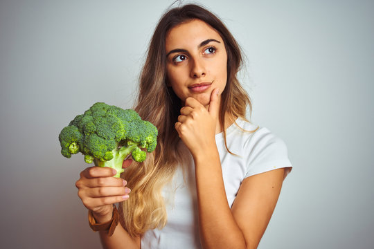 Young beautiful woman eating broccoli over grey isolated background serious face thinking about question, very confused idea