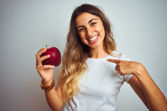 Young beautiful woman eating red apple over grey isolated background with surprise face pointing finger to himself