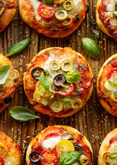 Mini pizzas with the addition of cherry tomatoes, olives, mozzarella cheese and fresh basil on a rustic wooden background, close-up, top view.