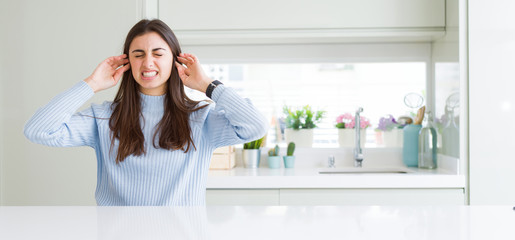 Wide angle picture of beautiful young woman sitting on white table at home covering ears with fingers with annoyed expression for the noise of loud music. Deaf concept.