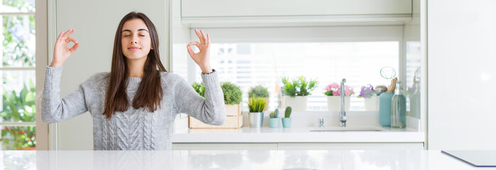 Wide angle picture of beautiful young woman sitting on white table at home relax and smiling with eyes closed doing meditation gesture with fingers. Yoga concept.