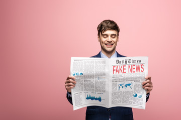 cheerful businessman reading newspaper with fake news and smiling on pink background