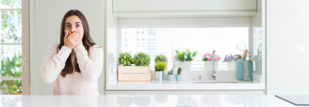 Wide angle picture of beautiful young woman sitting on white table at home shocked covering mouth with hands for mistake. Secret concept.