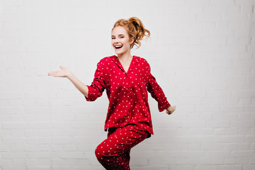 Slim positive caucasian girl in trendy red night-wear dancing on bricked background. Indoor photo...