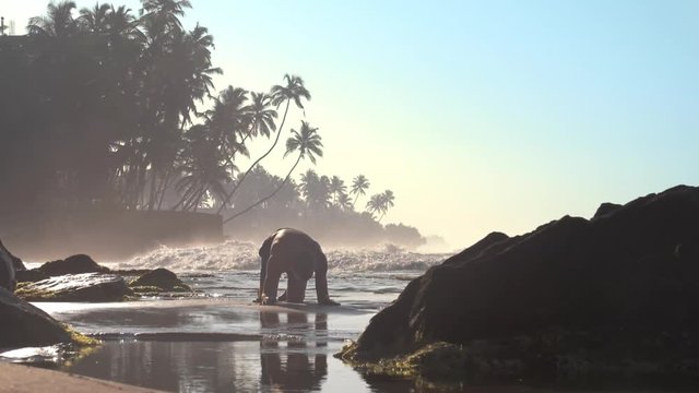 professional sportsman silhouette does difficult yoga exercise on wet sand against palms and ocean waves slow motion. Concept spiritual practics and meditation