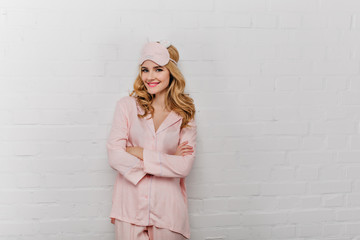 Joyful curly woman in silk pajama standing in confident pose near bricked wall. Positive lady in eyemask smiling on white background.