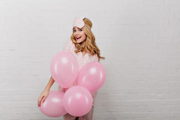 Surprised cheerful girl in sleepmask standing on white background with party balloons. Portrait of beautiful young lady celebrating something in weekend morning.