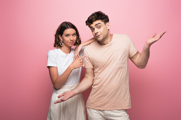 positive man showing shrug gesture while standing near smiling girlfriend on pink background