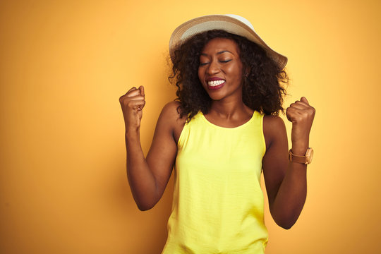 Young african american woman wearing t-shirt and hat over isolated yellow background very happy and excited doing winner gesture with arms raised, smiling and screaming for success. 
