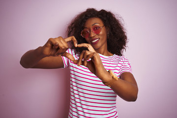 Young african american woman wearing t-shirt and sunglasses over isolated pink background smiling in love showing heart symbol and shape with hands. Romantic concept.