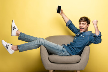 excited young man showing winner gesture while sitting in armchair and holding smartphone with...