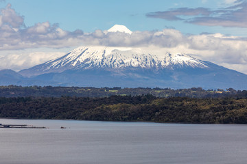 The amazing Osorno Volcano above the waters and clouds on a view from Rupanco Lake, the tress inside the forest and the clouds making it an awesome volcanic landscape. Perfect cone snow capped summit
