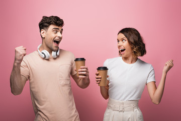 happy man and woman showing yes gestures while holding paper cups and looking at each other on pink background