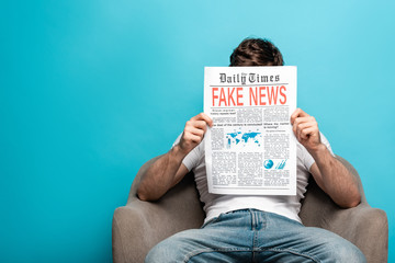 young man reading newspaper with fake news while sitting in armchair on blue background