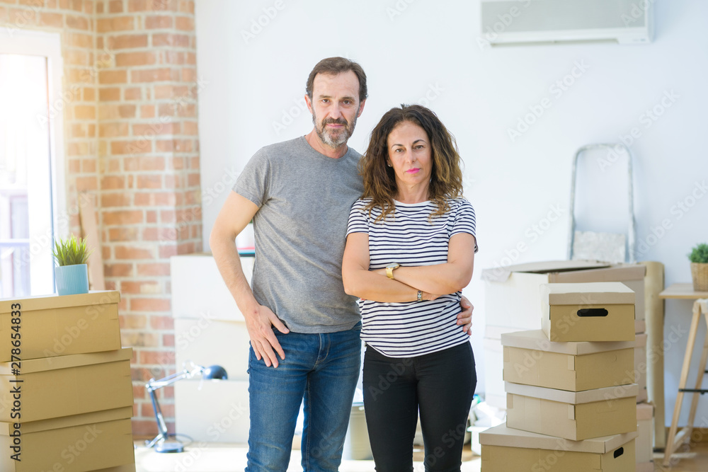 Poster Middle age senior couple moving to a new home with boxes around with serious expression on face. Simple and natural looking at the camera.