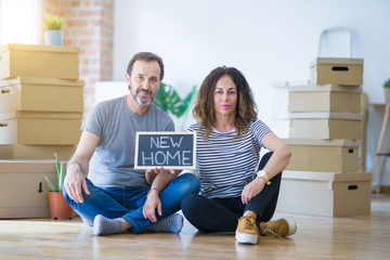 Middle age senior couple sitting on the floor holding blackboard moving to a new home with a...