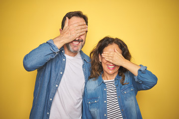 Beautiful middle age couple together wearing denim shirt over isolated yellow background smiling and laughing with hand on face covering eyes for surprise. Blind concept.