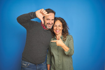 Beautiful middle age couple together standing over isolated blue background smiling making frame with hands and fingers with happy face. Creativity and photography concept.