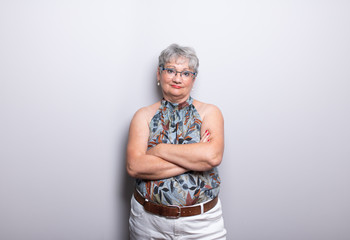 Elderly Woman with short gray hair and glasses posing in front of light gray wall with arms crossed wearing a self made neckholder top. the mature woman is looking at camera